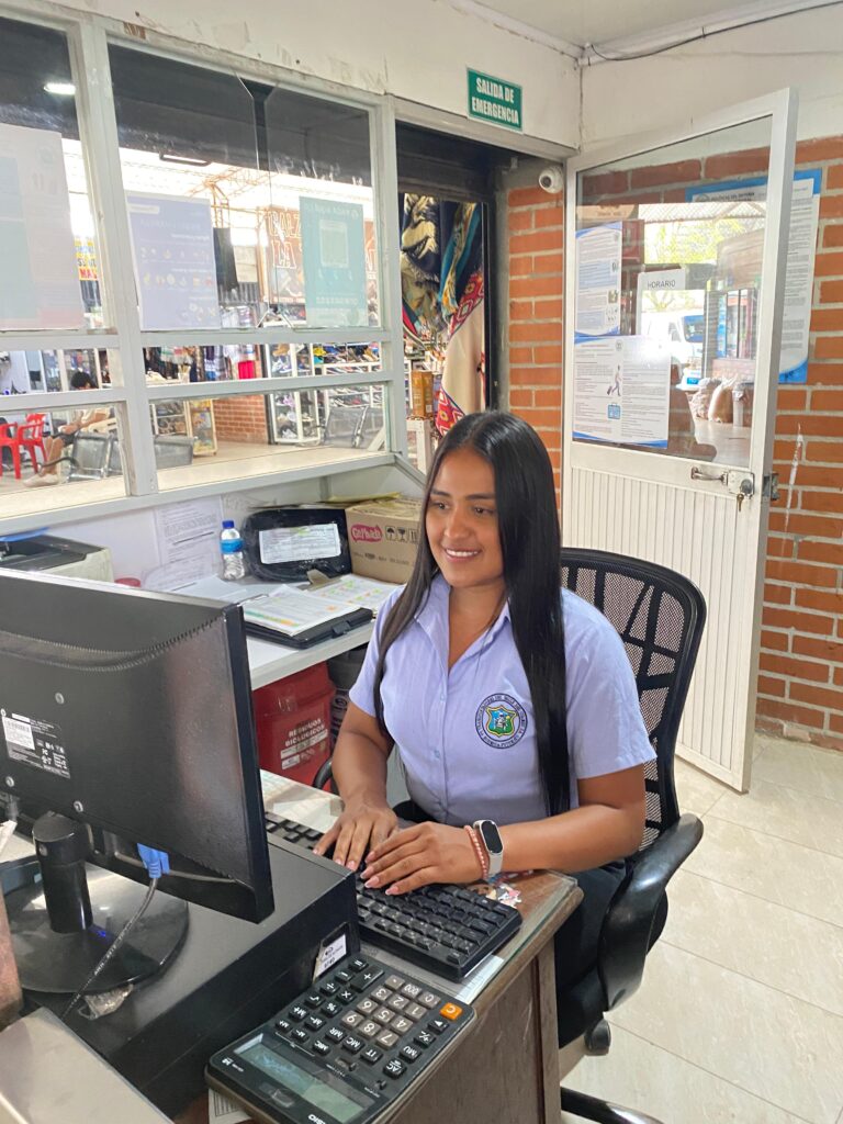 Empleada de atención al cliente en oficina, sonriendo mientras trabaja en el computador, lista para coordinar y ofrecer información sobre las rutas de transporte de la empresa.